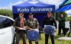 przed niebieskim namiotem przy białym samochodzie stoi czterech mężczyzn i trzymają w rękach opakowane siatki na ryby