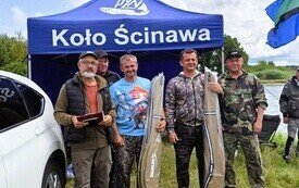 przed niebieskim namiotem przy białym samochodzie stoi czterech mężczyzn i trzymają w rękach opakowane pokrowce na wędki