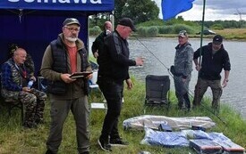przed niebieskim namiotem stoi mężczyzna a przed nim leżą na ziemi na trawie nagrody dla uczestnik&oacute;w zawod&oacute;w wędkarskich