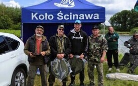 przed niebieskim namiotem przy białym samochodzie stoi czterech mężczyzn i trzymają w rękach opakowane siatki na ryby 