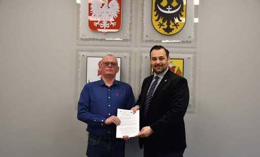 dwóch mężczyzn stoi obok siebie i razem trzymają w rękach dokument, w tle widoczne są cztery herby